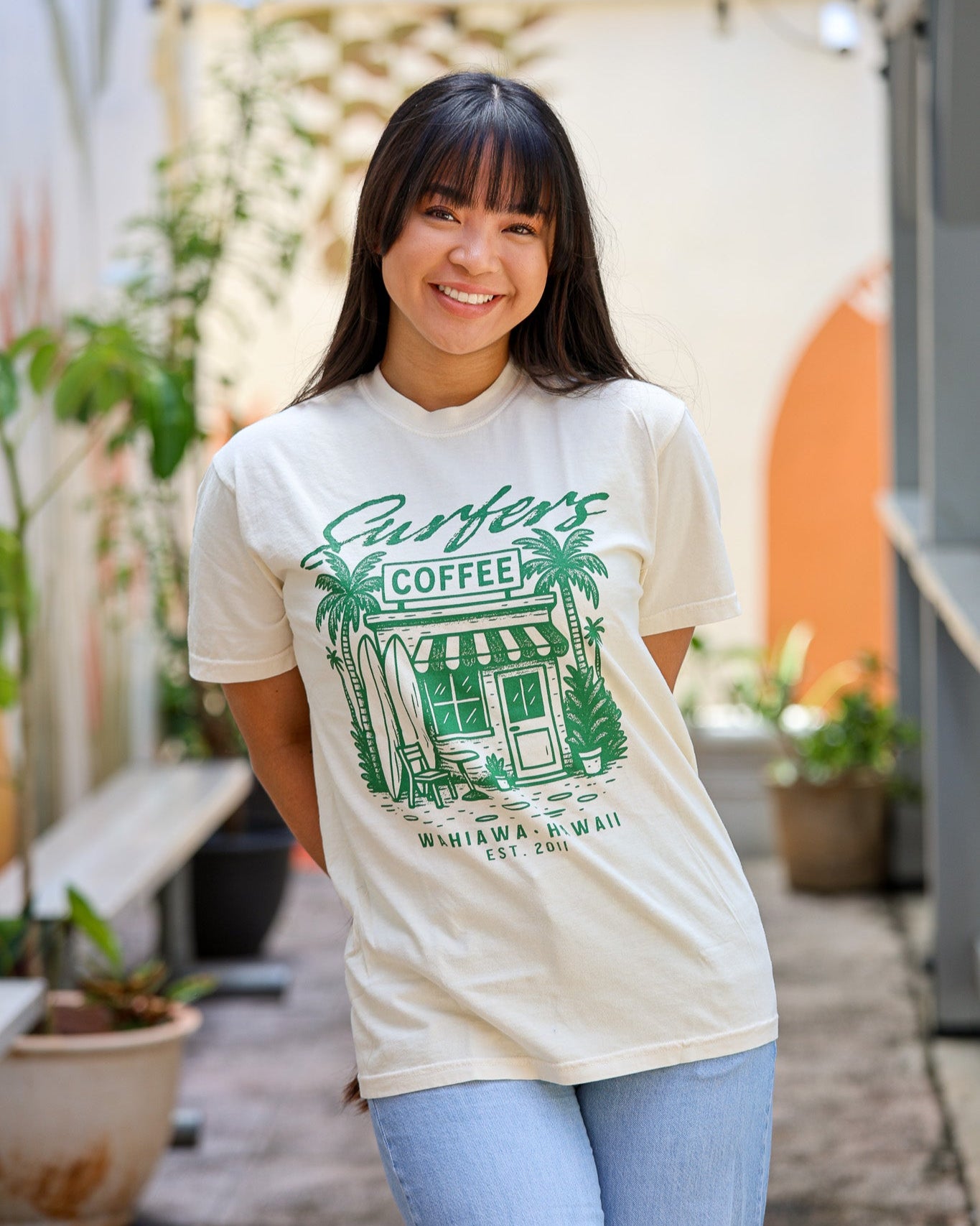 Woman wearing a t-shirt with a graphic design of a coffee shop and text, standing outdoors.