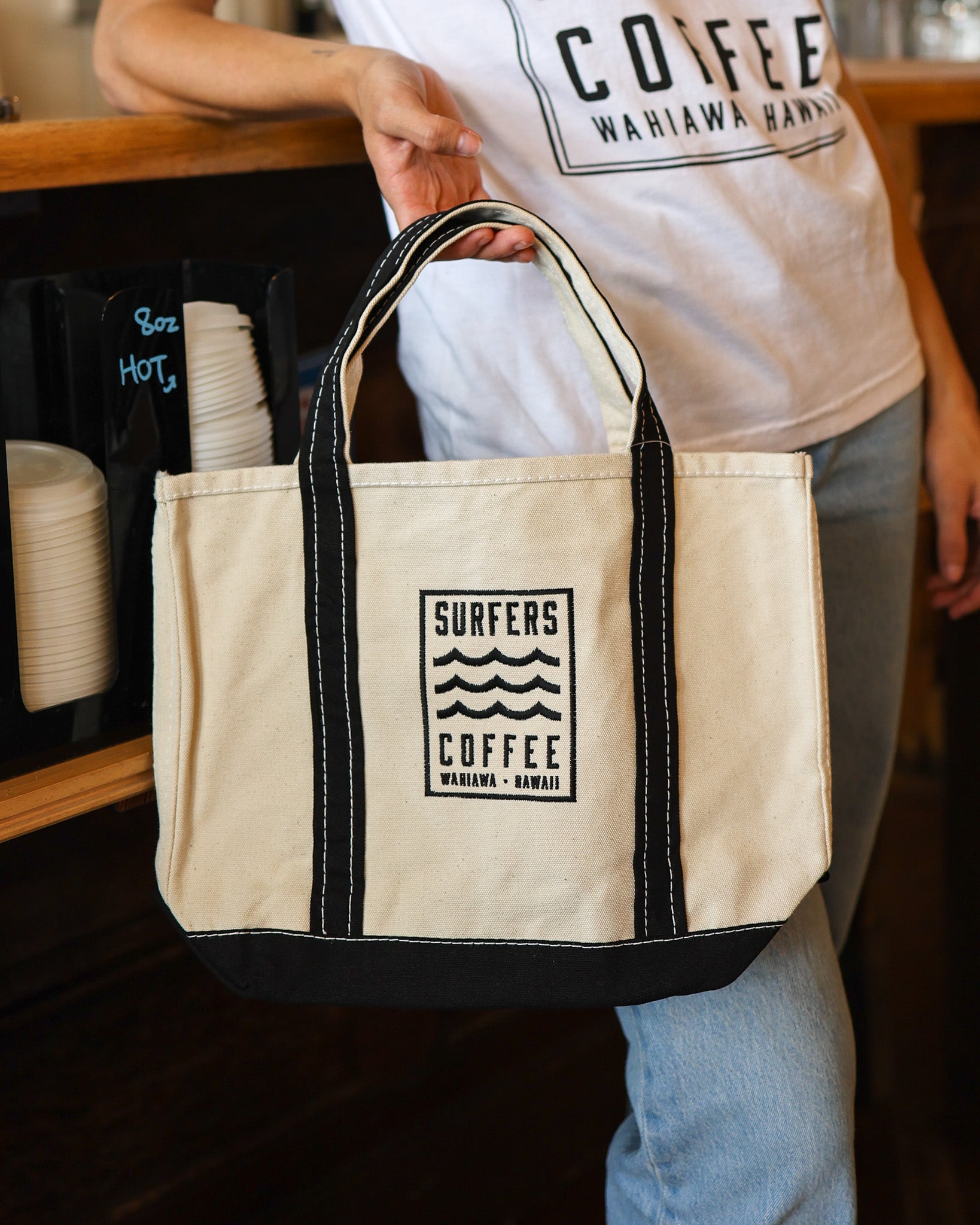 Beige tote bag with 'Surfers Coffee' logo held by a person wearing a 'Surfers Coffee' shirt.