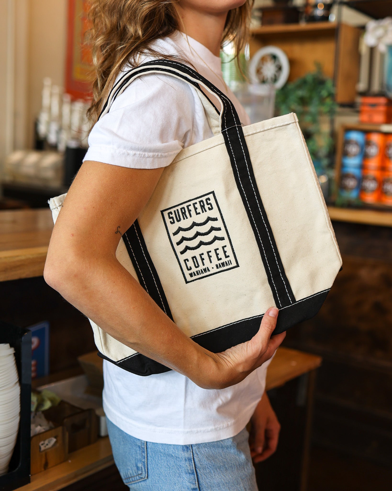 Person holding a beige tote bag with 'Surfers Coffee' logo in a store setting