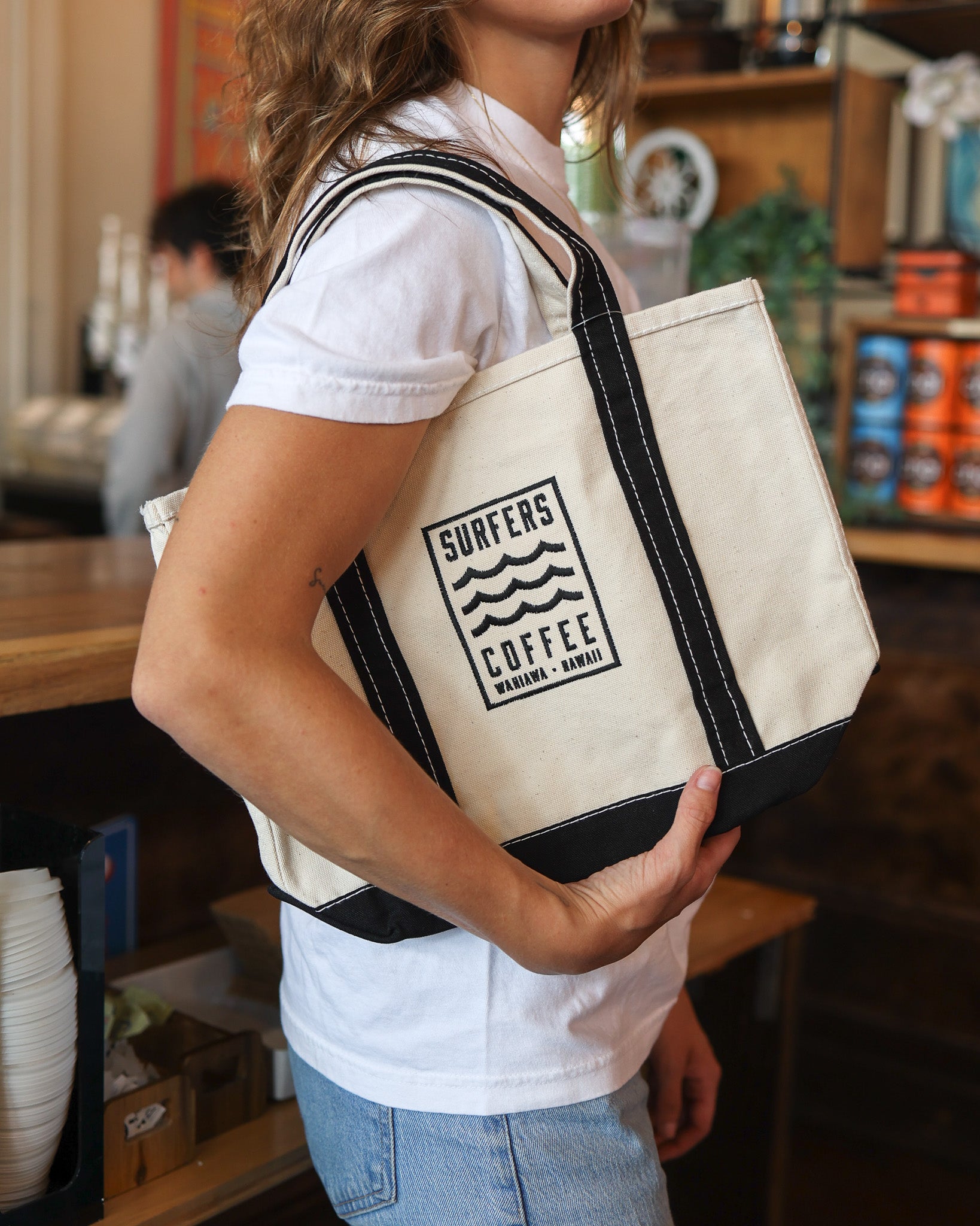 Person holding a beige tote bag with 'Surfers Coffee' logo in a casual setting.