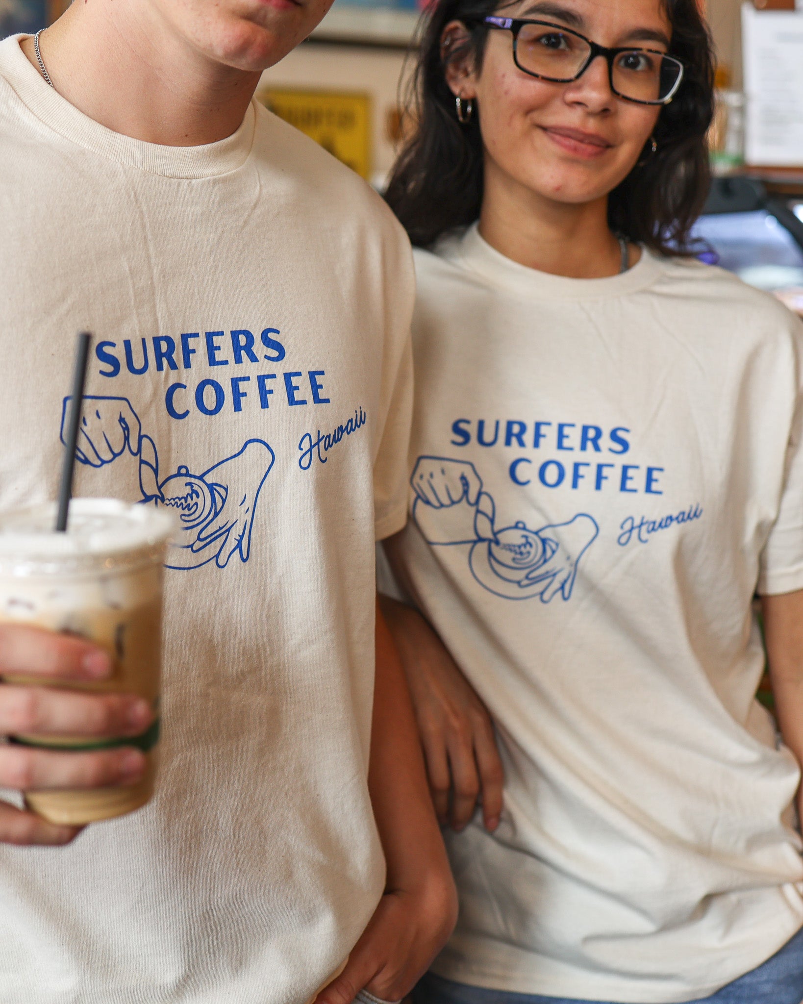 Two people wearing 'Surfers Coffee Hawaii' t-shirts, one holding a drink.