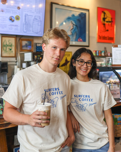 Two people wearing 'Surfers Coffee' shirts with a latte art graphic in a coffee shop setting.