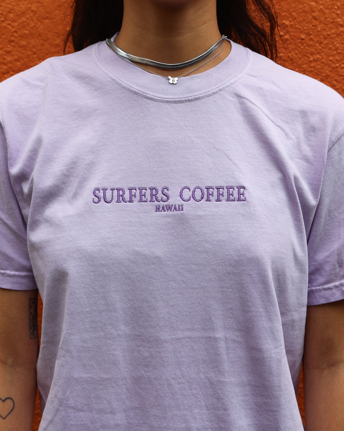 Person wearing a light purple t-shirt with 'Surfers Coffee Hawaii' text against an orange wall.